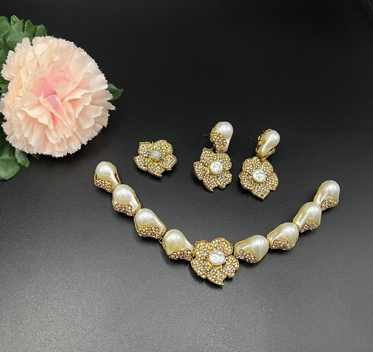 Gold necklace and earrings set with pearls on a dark surface, accompanied by a pink flower.